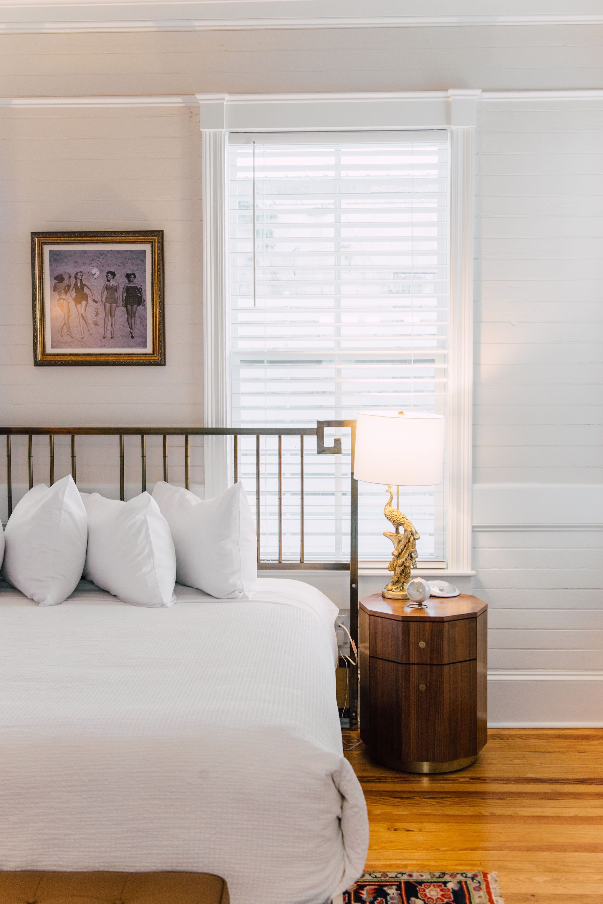 A neatly made bed with white linens and a headboard, a round wooden nightstand with a lamp, and a framed picture on the wall.