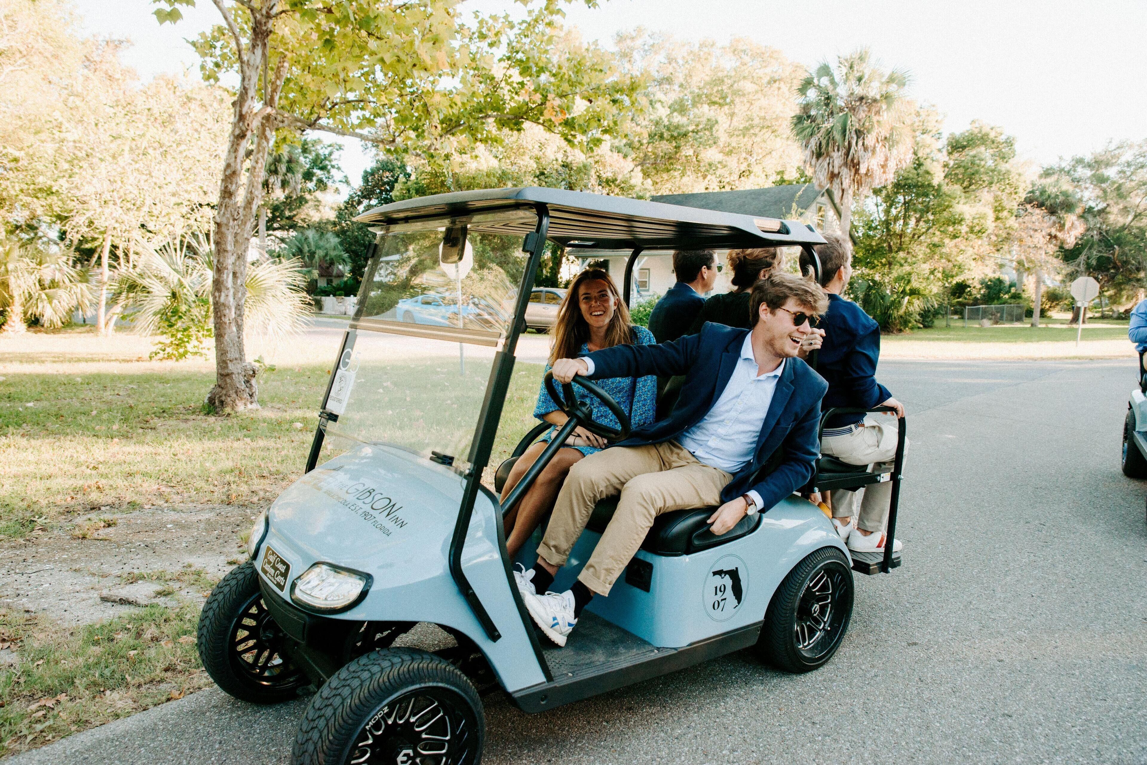 A golf cart with passengers, parked on a street with trees and a house in the background.