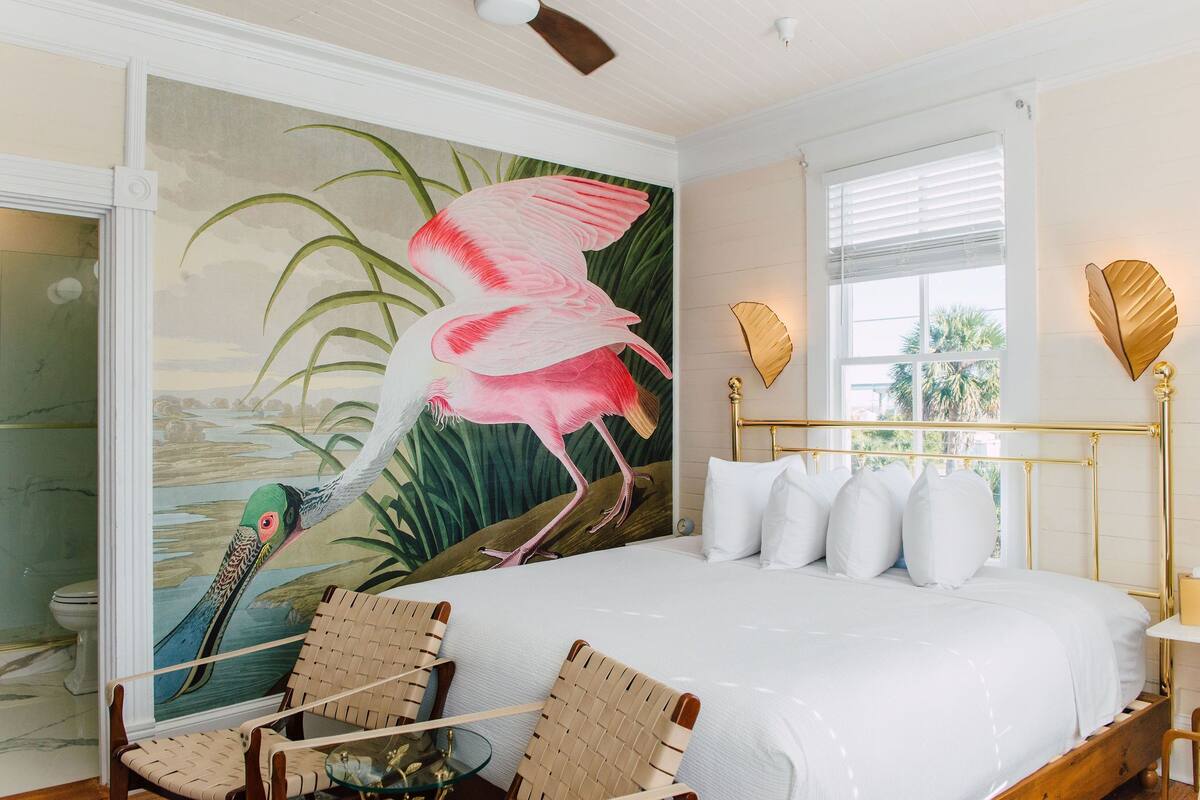 A bedroom with a large bed, a mural of a flamingo, two wicker chairs, and a window with blinds.