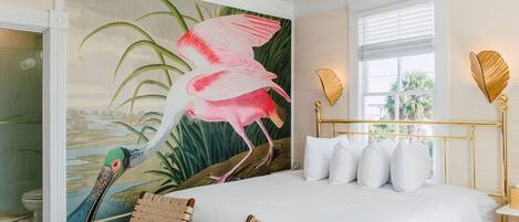 Un dormitorio con una cama grande, un mural de un flamenco, dos sillas de mimbre y una ventana con persianas.