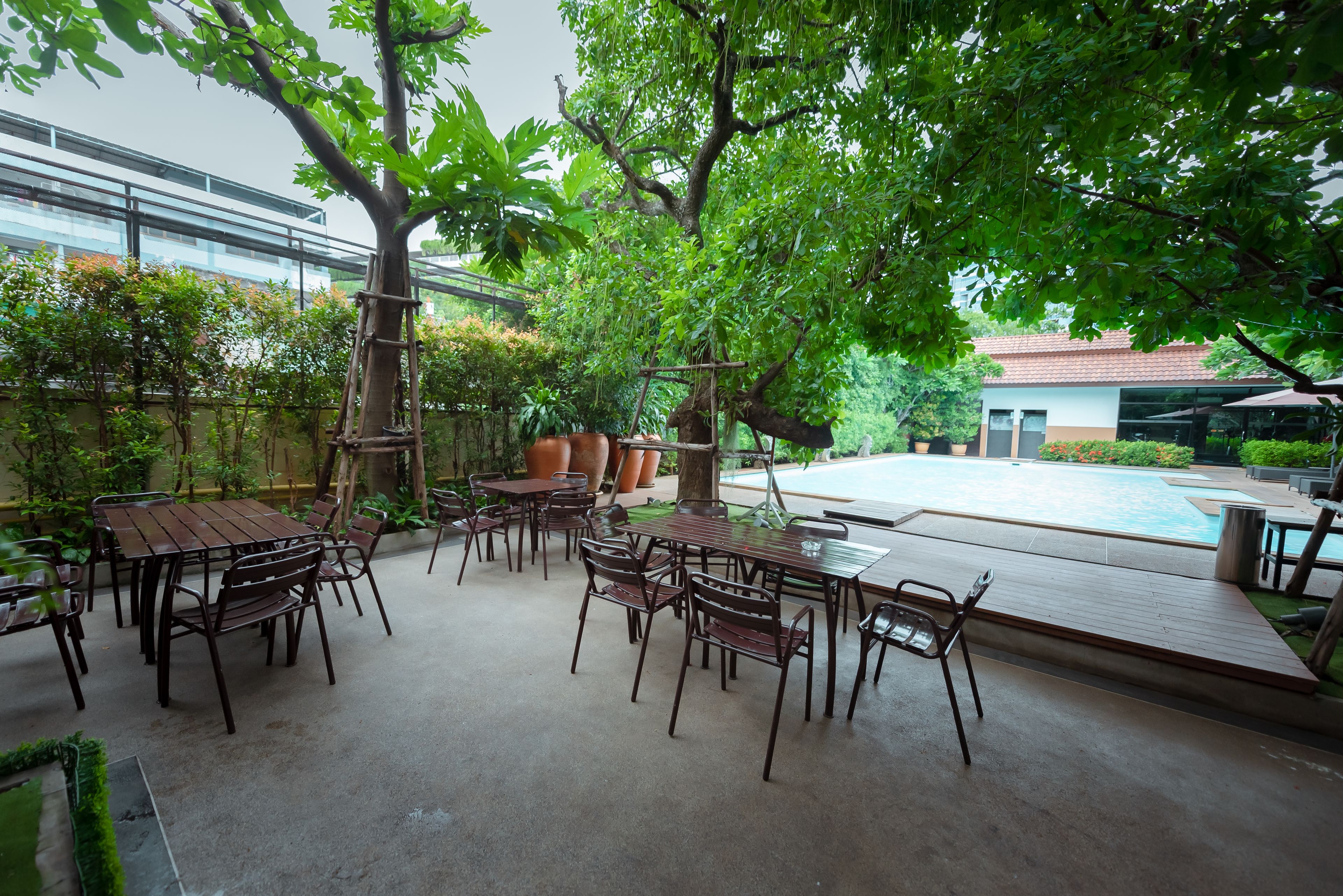 An outdoor patio area with wooden tables and chairs, surrounded by lush greenery and a swimming pool.