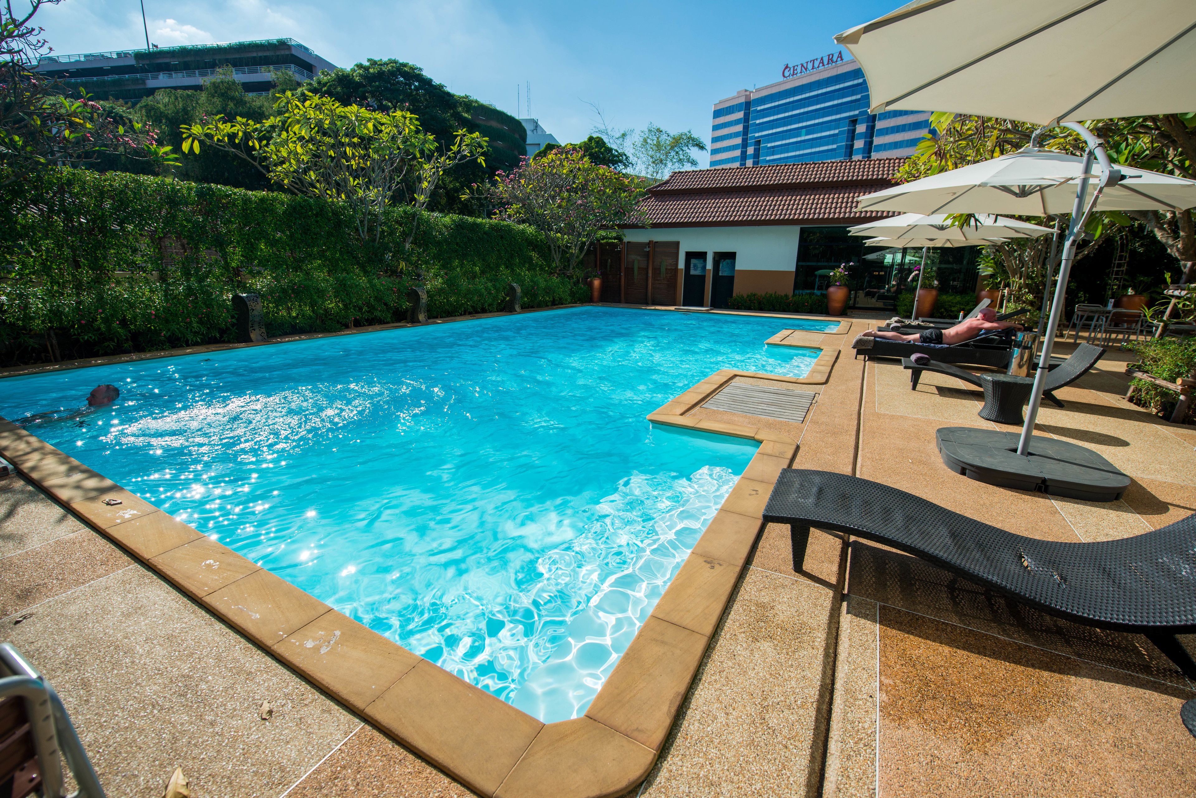 Una piscina con sillones de descanso y sombrillas, rodeada de vegetación y un edificio al fondo.