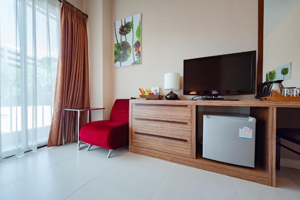 A hotel room with a wooden desk, a television, a red chair, a small refrigerator, and a window with curtains.