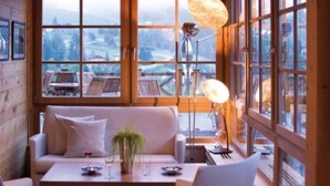 Ein gemütliches Blockhaus-Interieur mit einer Couch, zwei Sesseln, einem Esstisch und großen Fenstern mit Blick auf eine verschneite Landschaft.