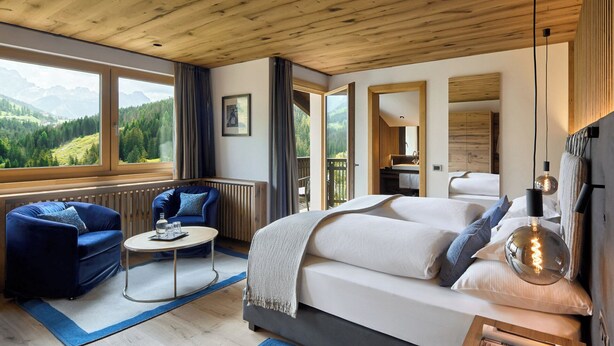 A modern hotel room with a large bed, a seating area with a blue sofa, a small round table, and a view of the mountains. - La Majun (Badia)