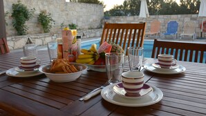 A dining table set with plates, cups, and a bowl of snacks, overlooking a pool area with lounge chairs and umbrellas.