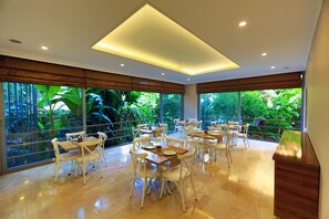 A spacious dining area with multiple tables and chairs, large windows offering a view of lush greenery, and a modern ceiling design.