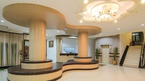 Um hall amplo com um balcão de recepção curvo, grandes colunas de madeira, um candelabro e uma escadaria.