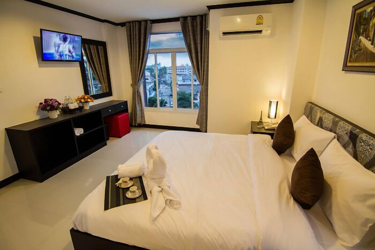 A hotel room with a large bed, a TV on the wall, a window with curtains, a desk with a lamp, and a view of the city.