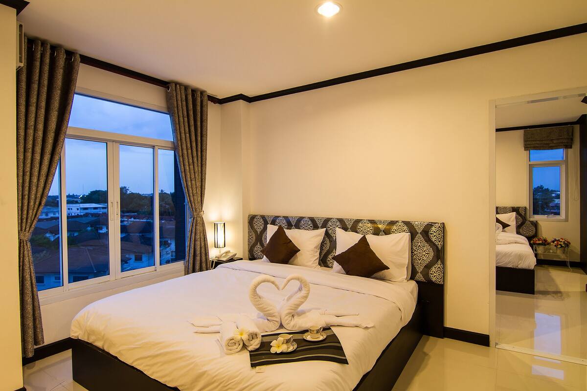 A hotel room with a large bed, a nightstand, a mirror, and a view of the cityscape through the window.