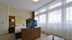Habitación de hotel con cama, televisor en un mueble de madera, mesita de noche y un florero con una flor sobre una mesa.
