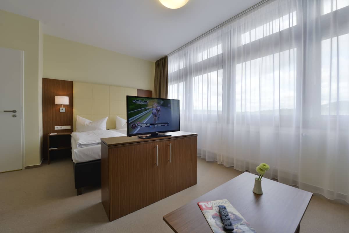 A hotel room with a bed, a TV on a wooden cabinet, a bedside table, a nightstand, a window with sheer curtains, and a vase with a flower on a table.