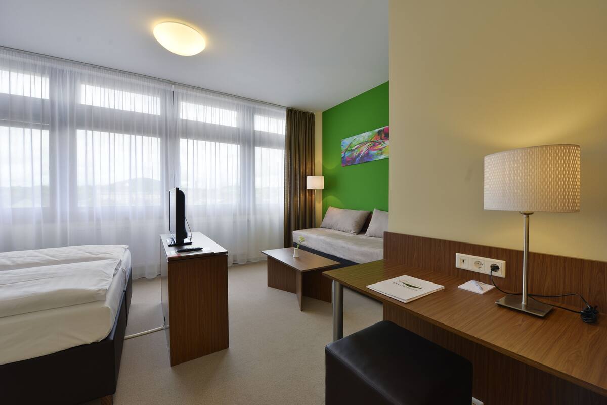 A hotel room with a bed, a desk, a chair, a lamp, and a painting on the wall.