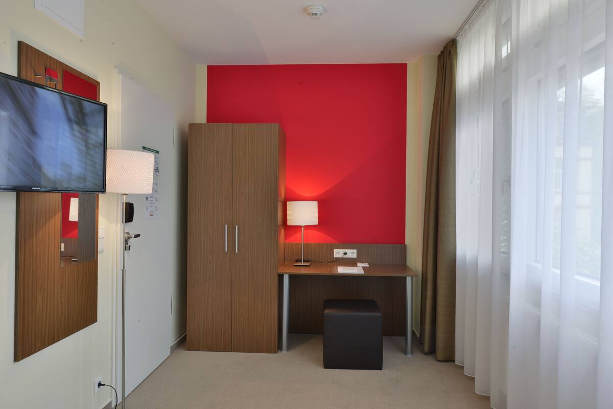 A hotel room with a red accent wall, a desk with a lamp, a television, and a wardrobe.
