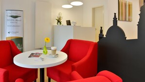 Zona de recepción de un hotel con sillas rojas, una mesa blanca con café y un jarrón de flores, y un mostrador de recepción.
