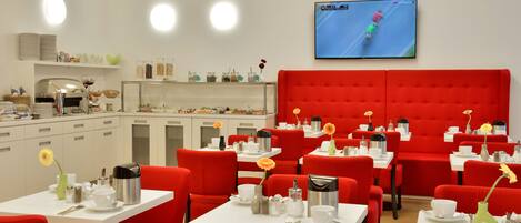 Un comedor moderno con sillas tapizadas en rojo y mesas blancas, una cocina al fondo, y un televisor mostrando una competición de carreras.