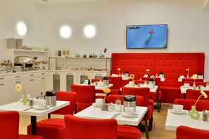 Un espace repas moderne avec des chaises à revêtement rouge et des tables blanches, une cuisine en arrière-plan et un téléviseur diffusant une course.