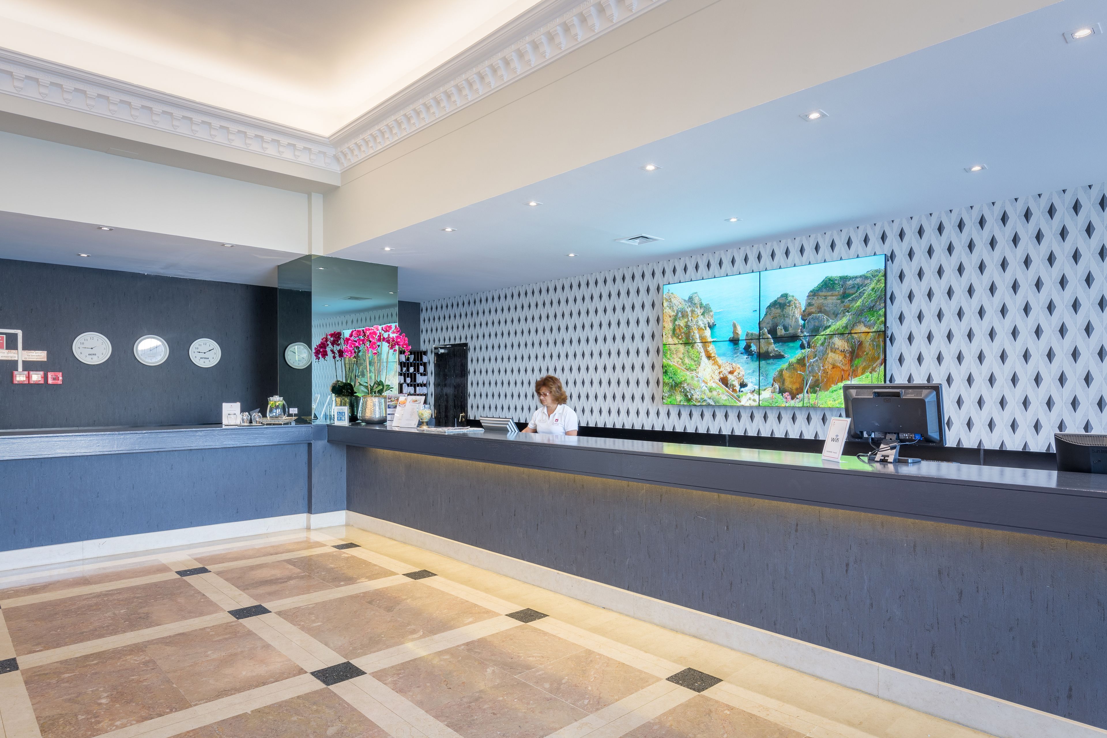 A hotel reception area with a modern design, featuring a large digital screen displaying a scenic view, a reception desk, and a check-in counter.