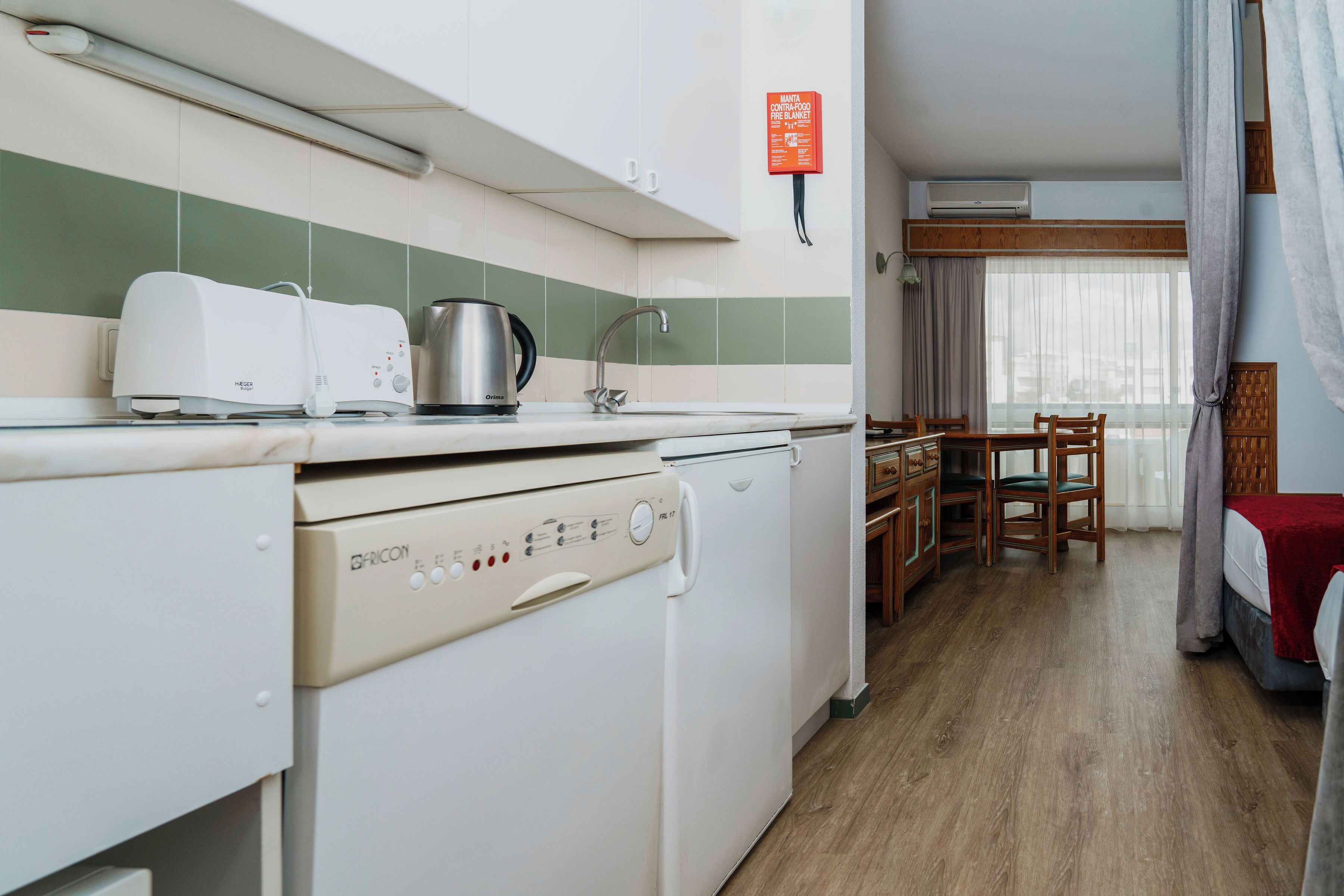 A compact kitchen with a toaster, kettle, and dishwasher, a dining area with a table and chairs, and a bed with a red blanket.