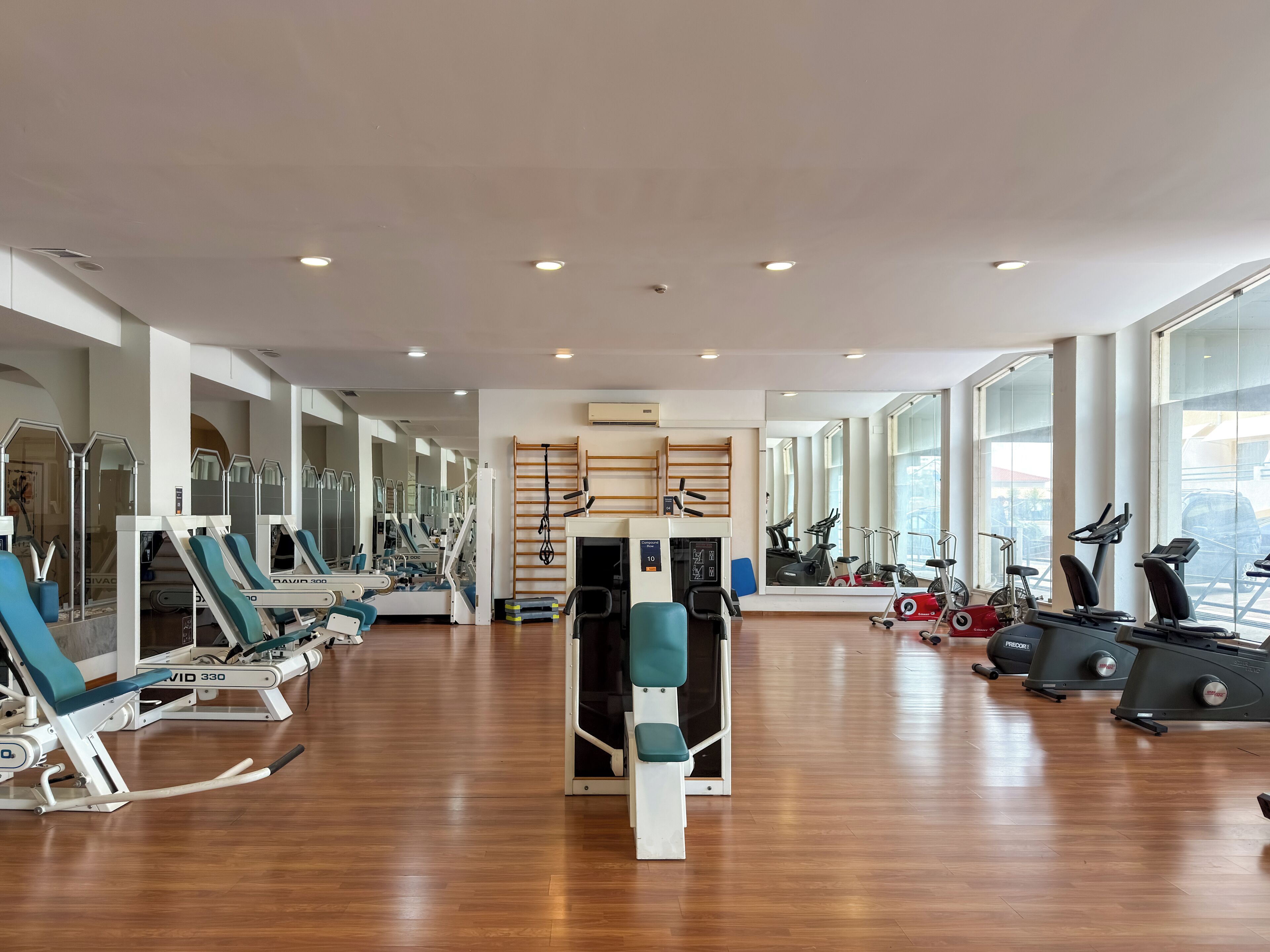 A spacious gym with various exercise machines, large windows, and a wooden floor.