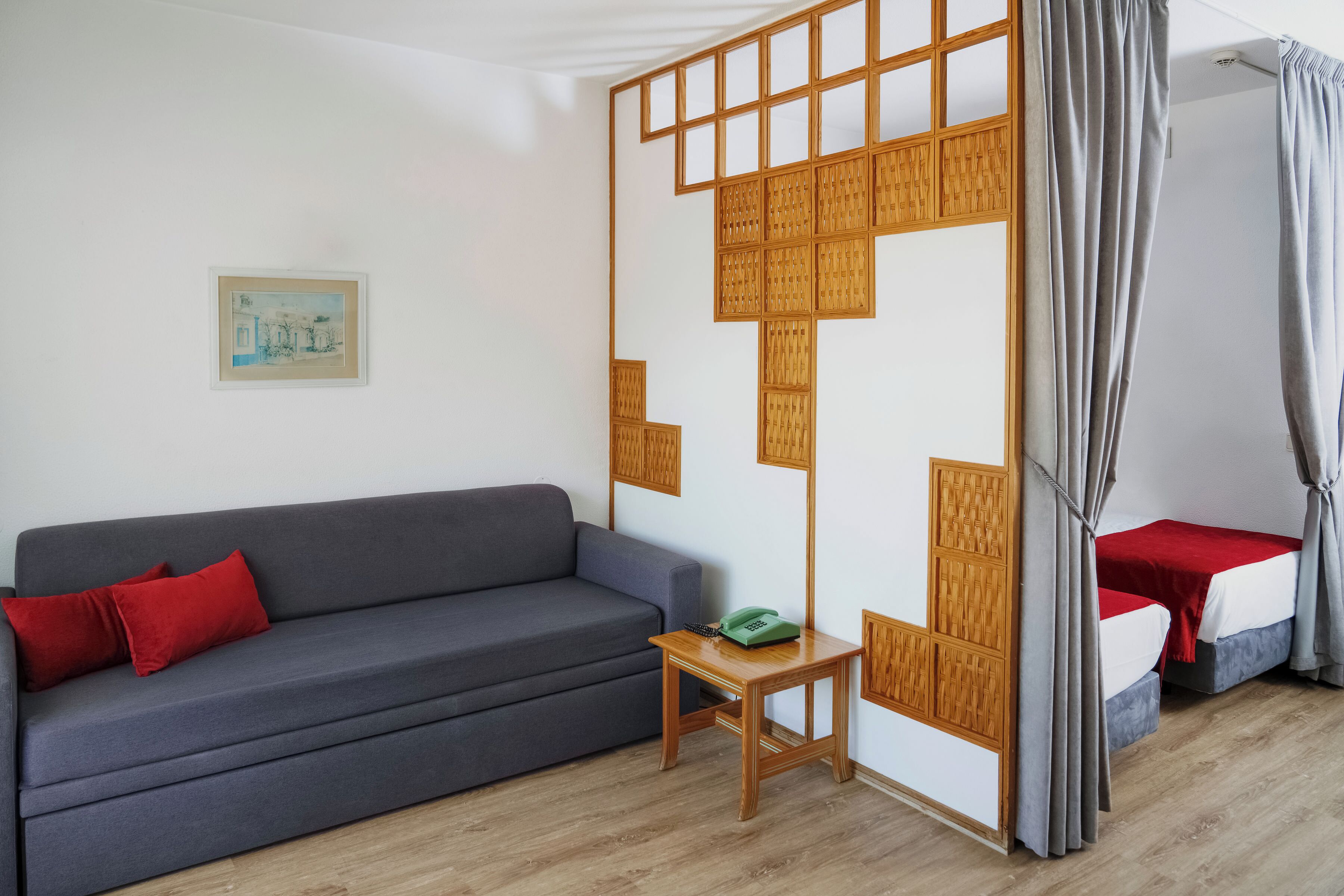 A room with a grey sofa, a small wooden table with a green telephone, a bed with grey curtains, and a wall-mounted wooden partition.