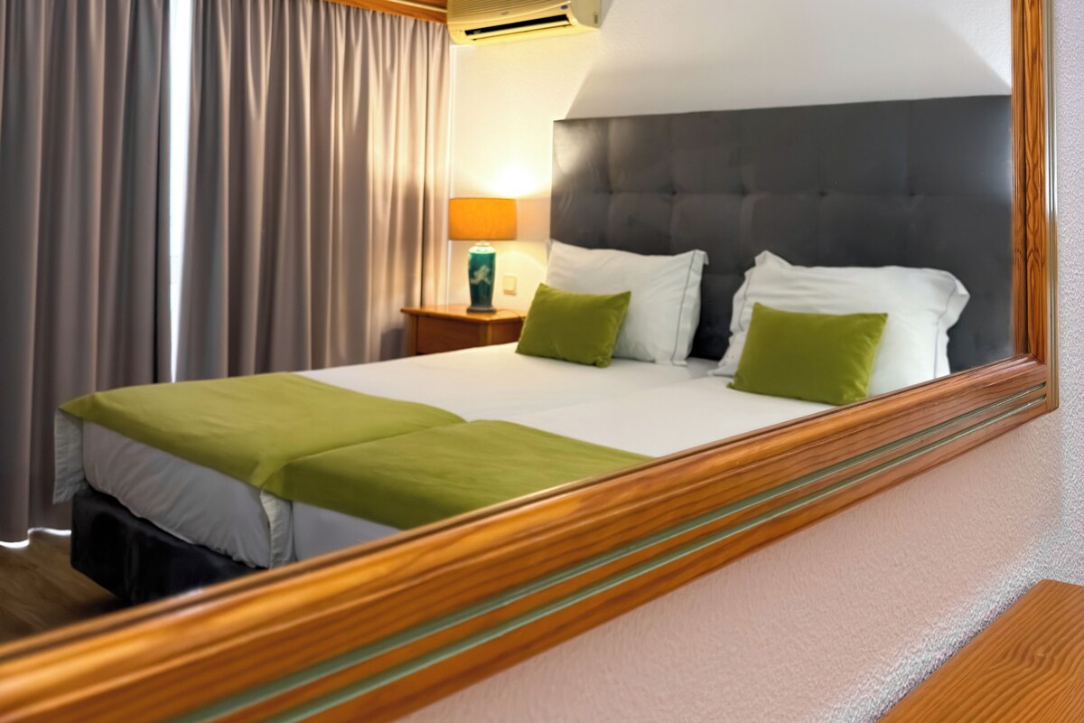A hotel room with a bed, a nightstand, a lamp, an air conditioning unit, and a large mirror.