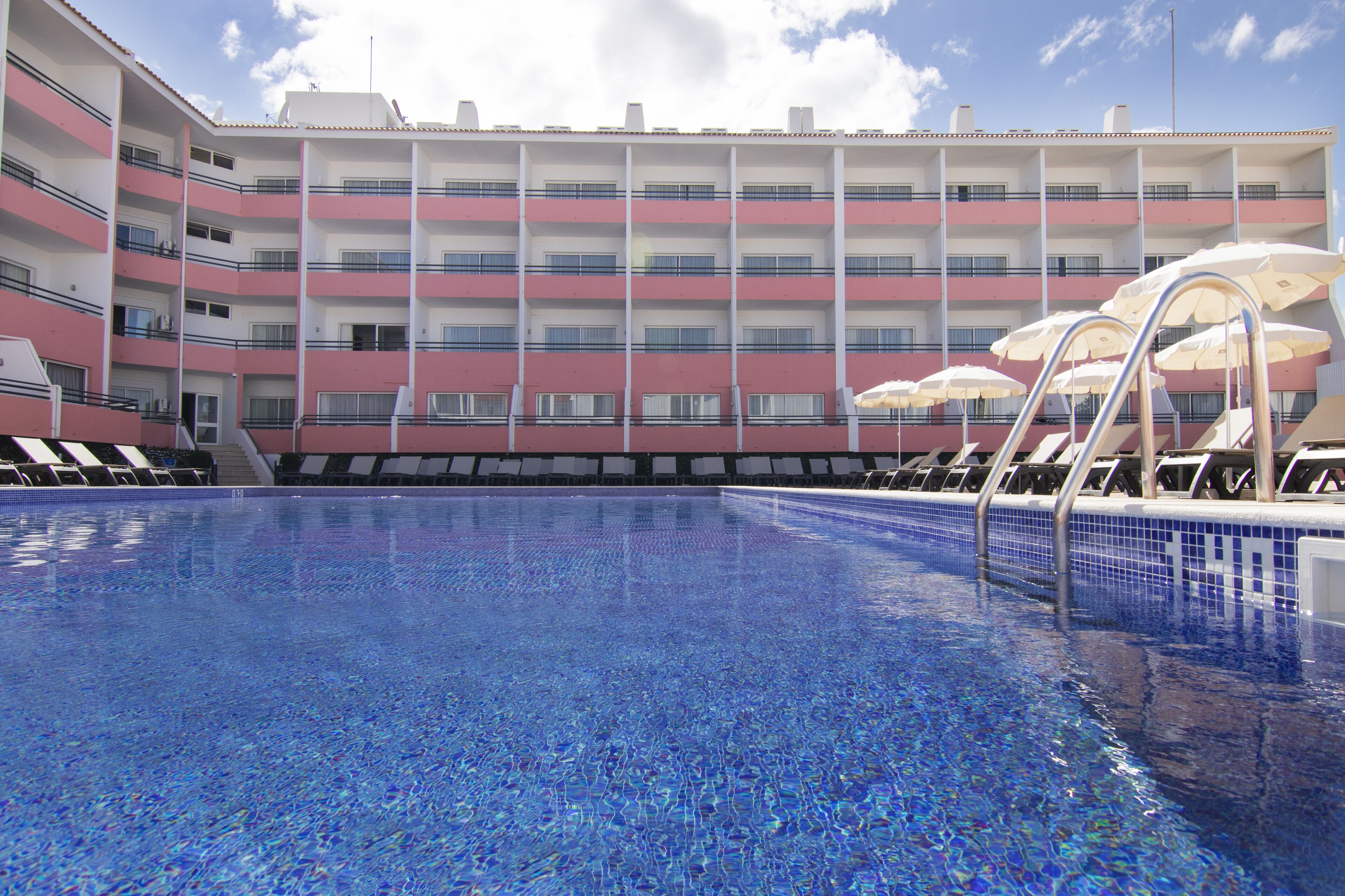 A swimming pool with a tiled floor, surrounded by a multi-story building with balconies and umbrellas.