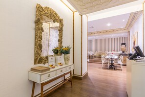 Zona de recepción de hotel con una consola blanca, un espejo grande y ornamentado, y un florero con flores. Hay un mostrador de recepción con una computadora y una silla. El cuarto tiene piso de madera y una pared rayada de fondo.