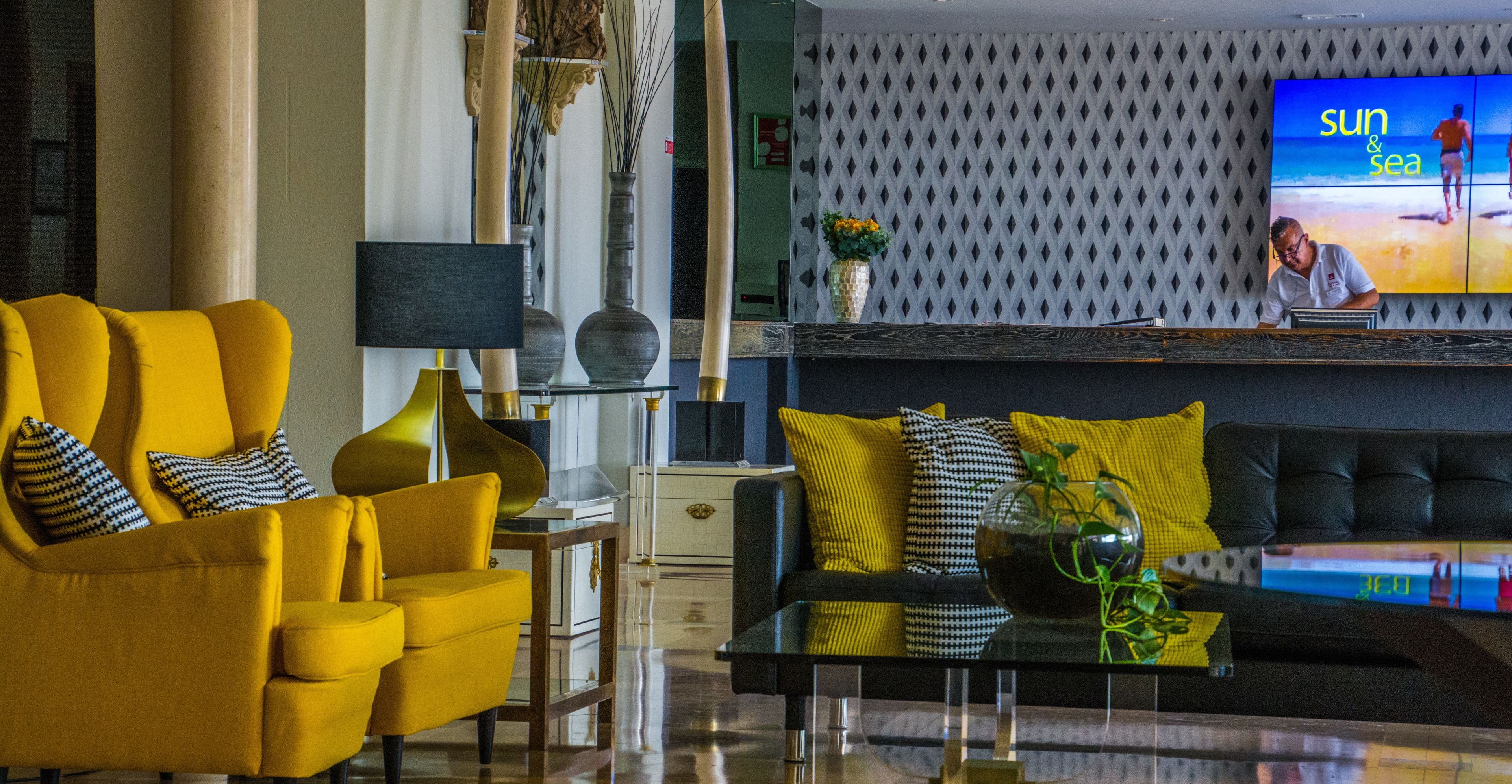 A modern living room with a yellow sofa, a black sofa, and a glass coffee table. There is a television on the wall displaying a beach scene.