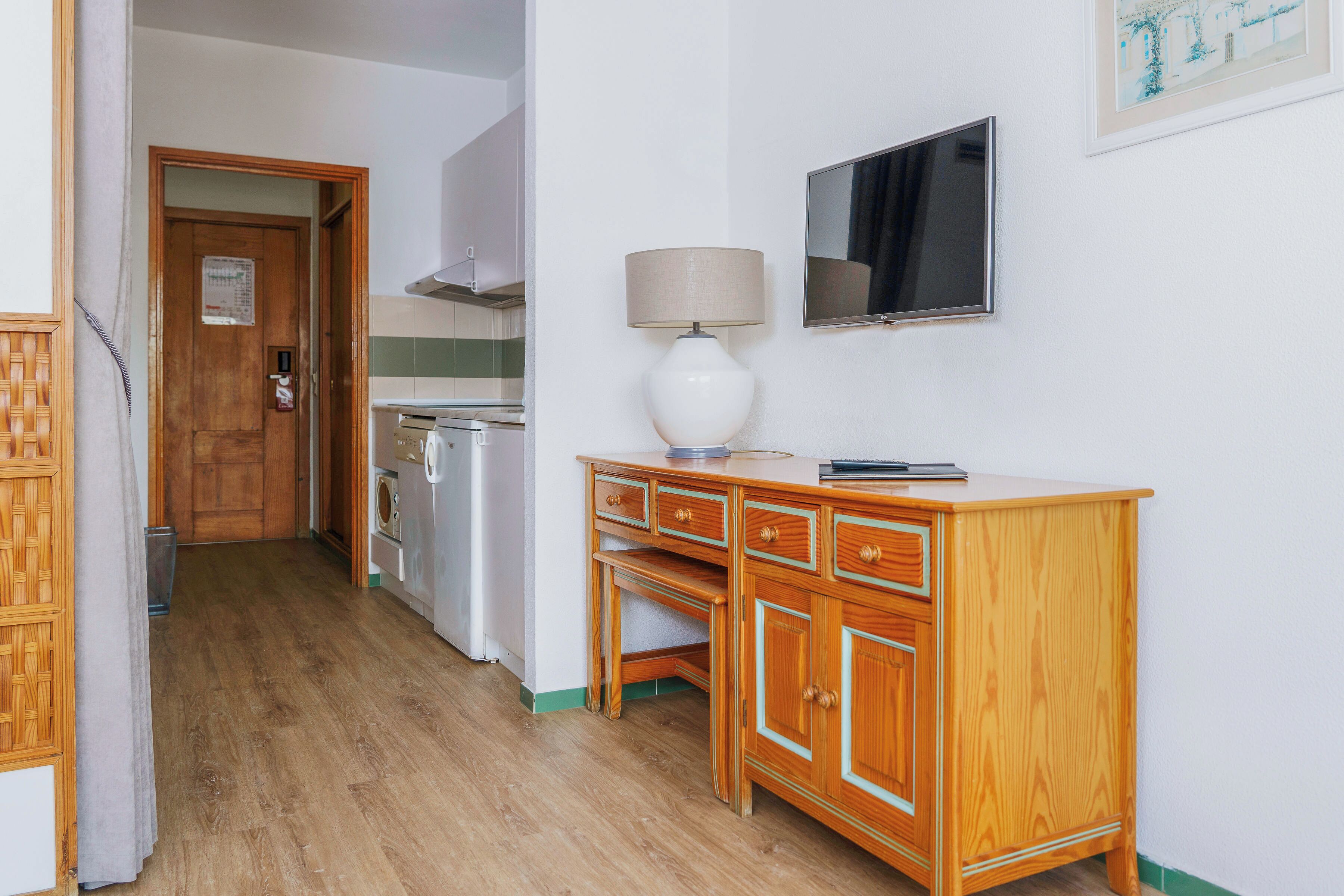 A hallway with a wooden desk, a lamp, a television, and a door leading to a kitchen area.