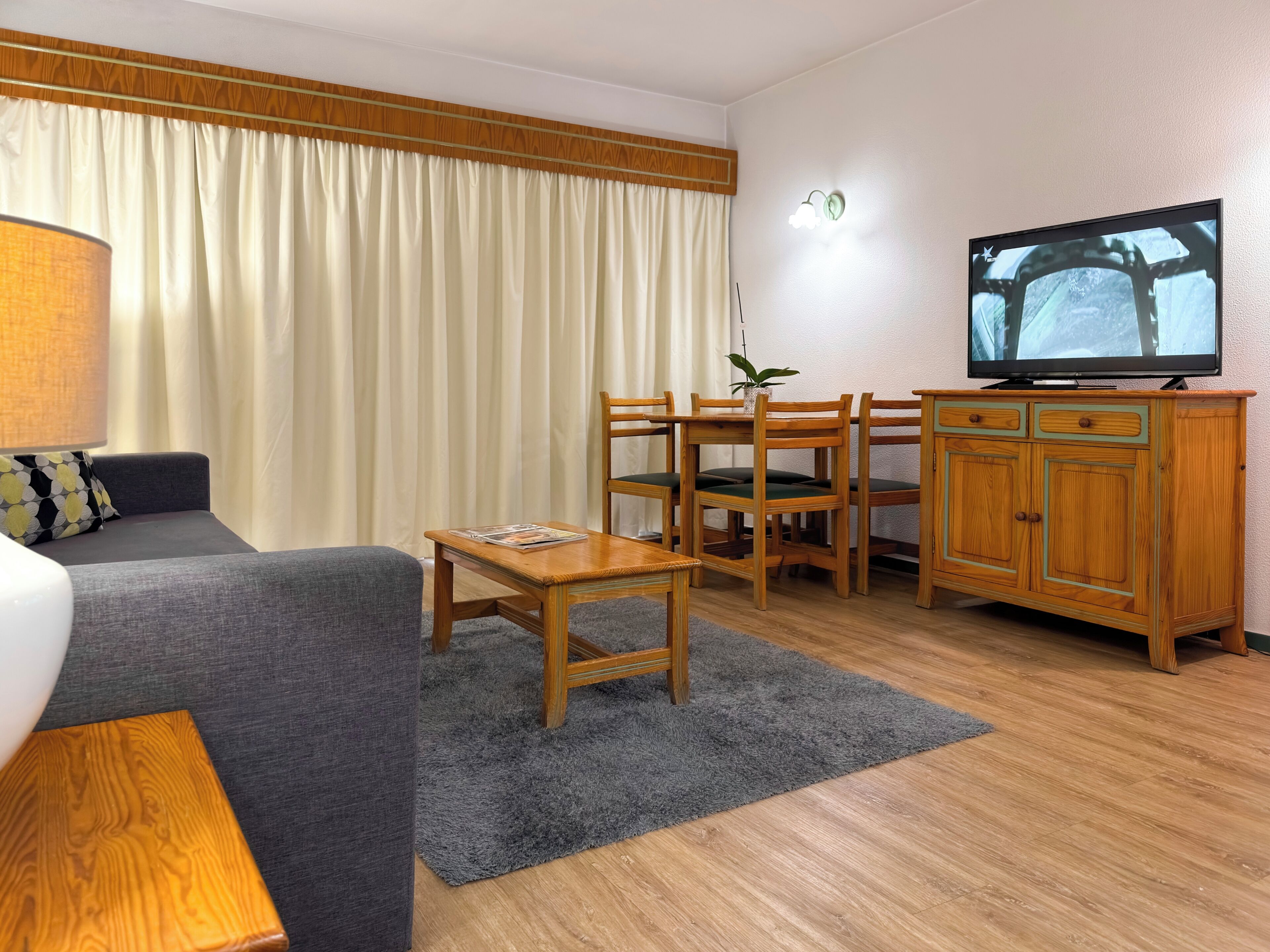 Una habitación de hotel con sofá, una mesita, televisor, aparador de madera y ventana con cortinas.