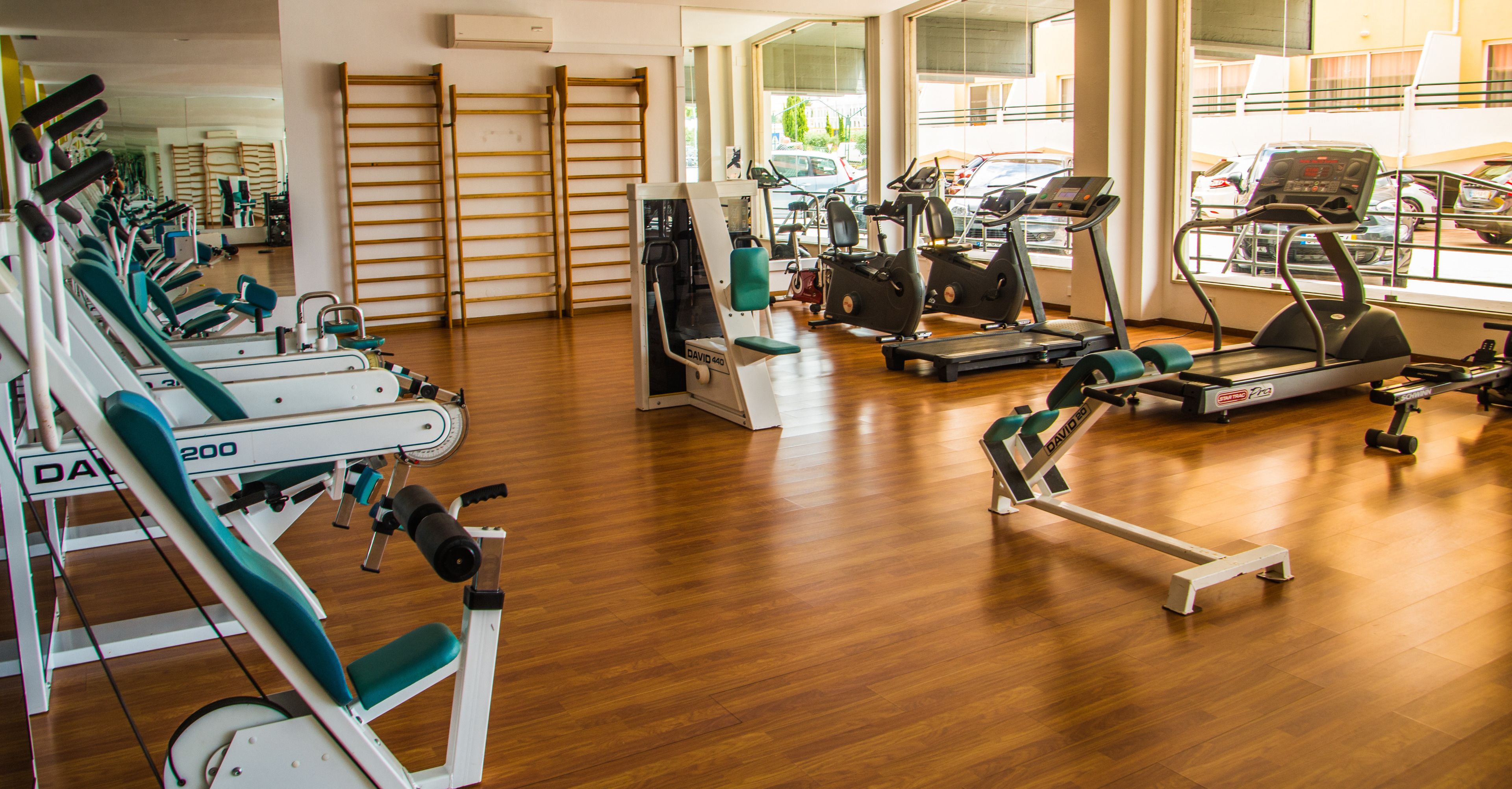 Un gimnasio con diversos equipos de ejercicio, incluyendo cintas de correr y máquinas de pesas, y amplias ventanas que ofrecen vistas a la calle.
