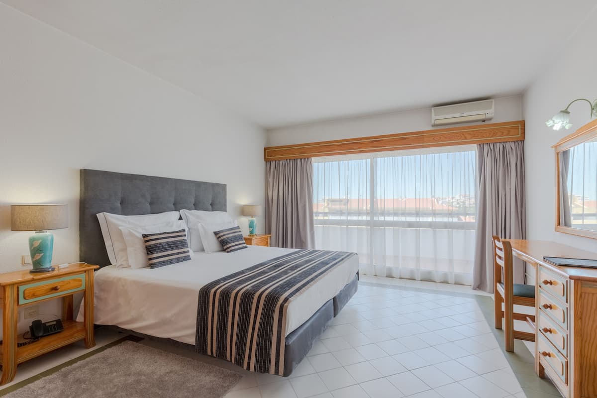A hotel room with a bed, a desk, a chair, a window with curtains, and an air conditioning unit.
