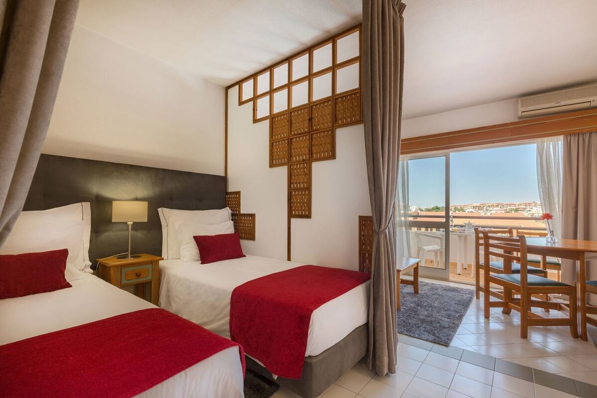 A hotel room with two beds, a balcony with a view, a dining area, and an air conditioning unit.