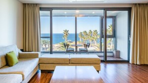 A spacious room with a large sliding glass door leading to a balcony with a view of the ocean and palm trees. There is a sofa and a small table in the room.