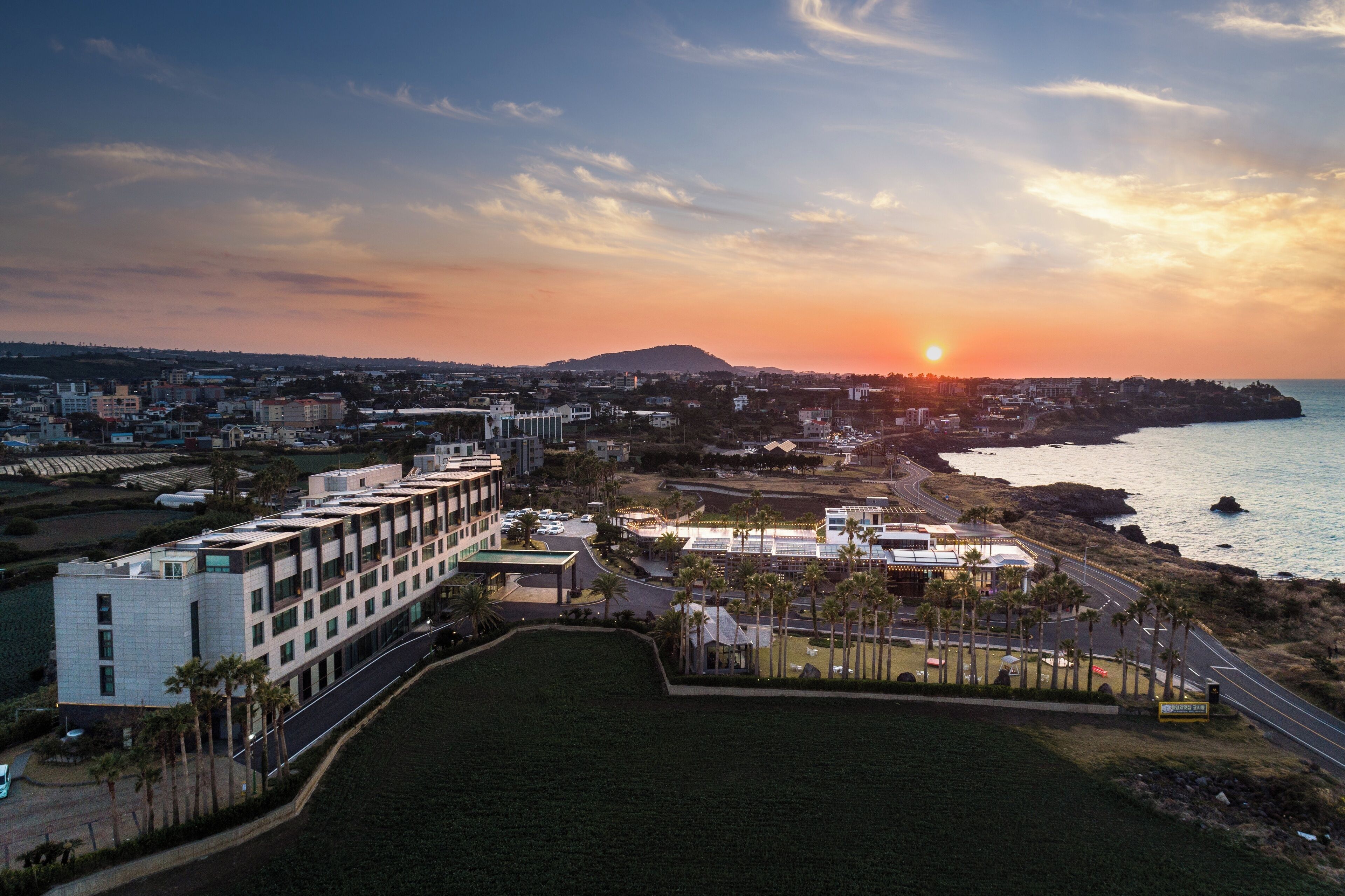 A coastal cityscape with a modern building complex, a marina, and a sunset over the ocean.