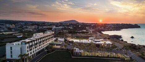 A coastal cityscape with a modern building complex, a marina, and a sunset over the ocean.