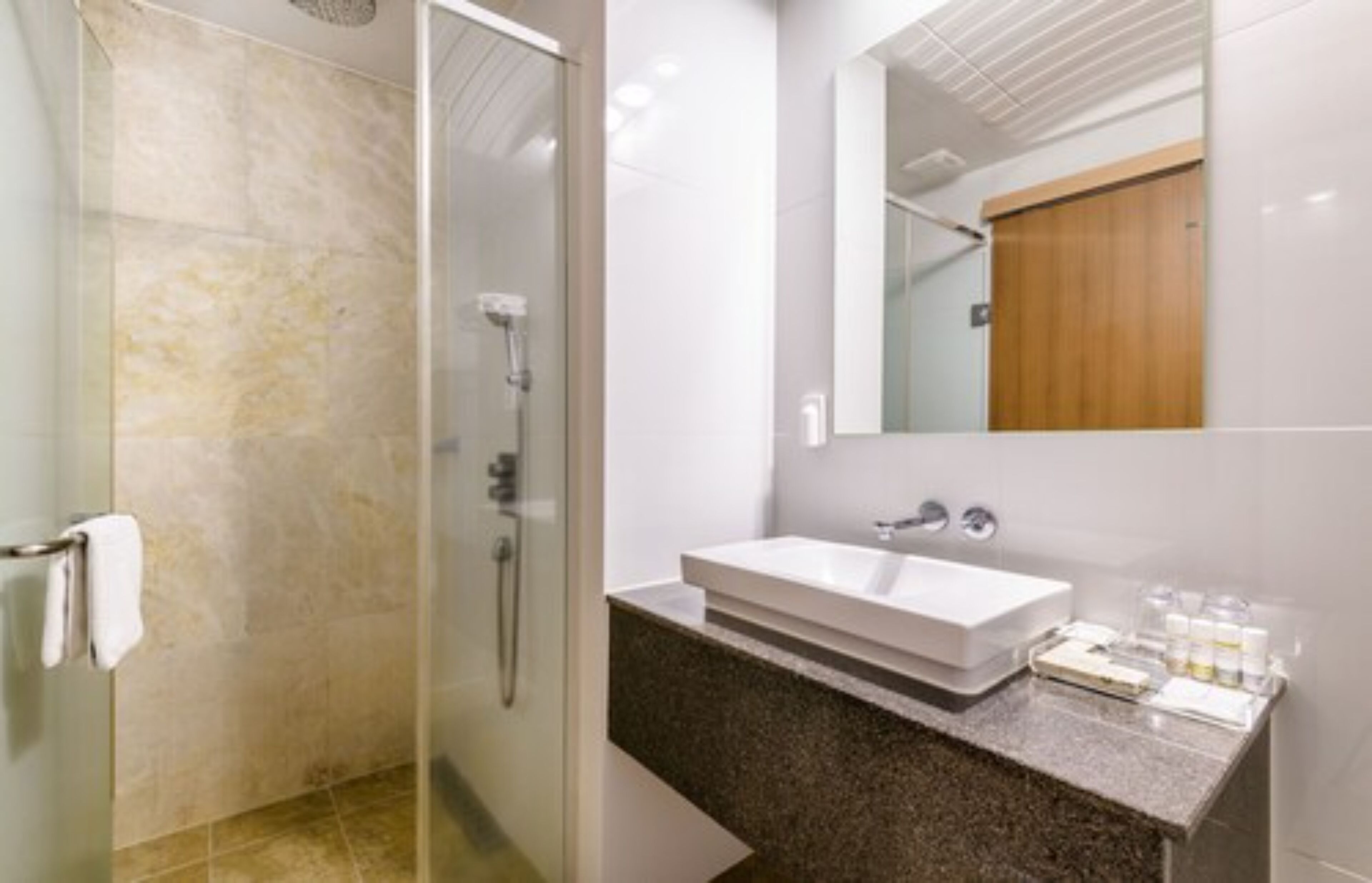 Un baño moderno con cabina de ducha de vidrio, un lavabo blanco con encimera de granito y una puerta de madera al fondo.
