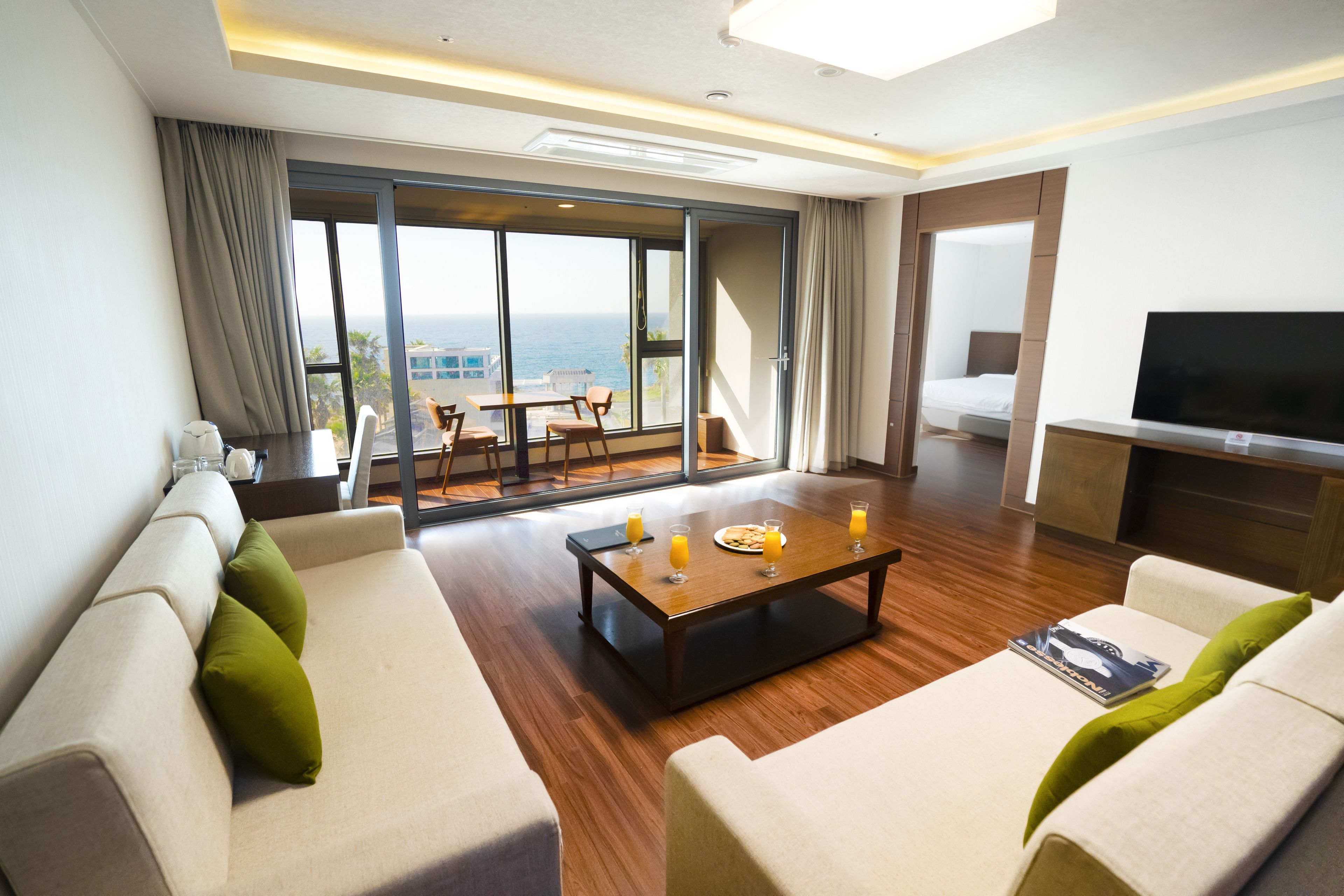 A modern hotel room with a large sliding glass door leading to a balcony with a view of the ocean. There are two sofas, a coffee table, and a flat-screen TV.