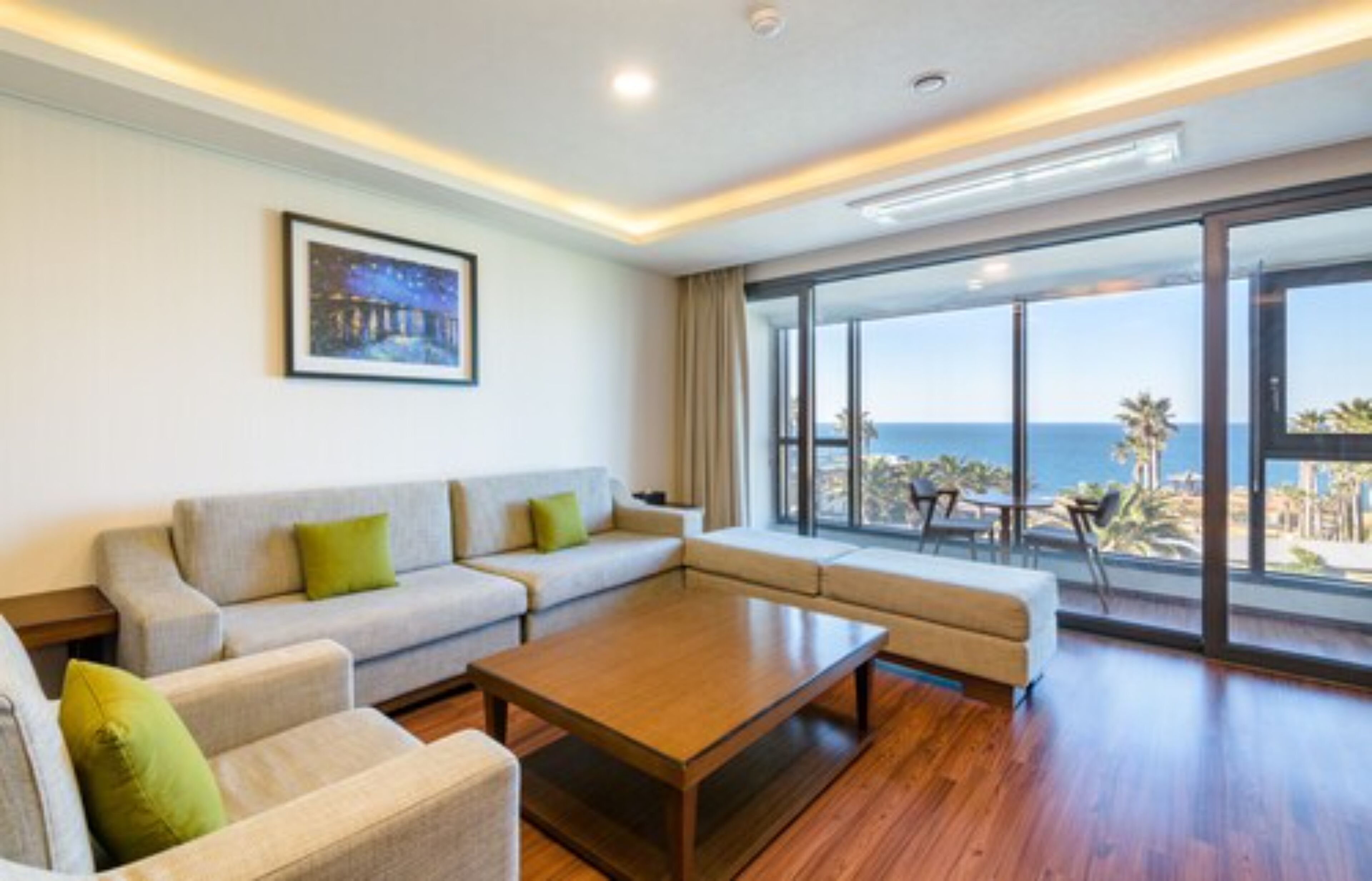 A modern living room with a sofa set, a coffee table, and large windows offering a view of the sea.