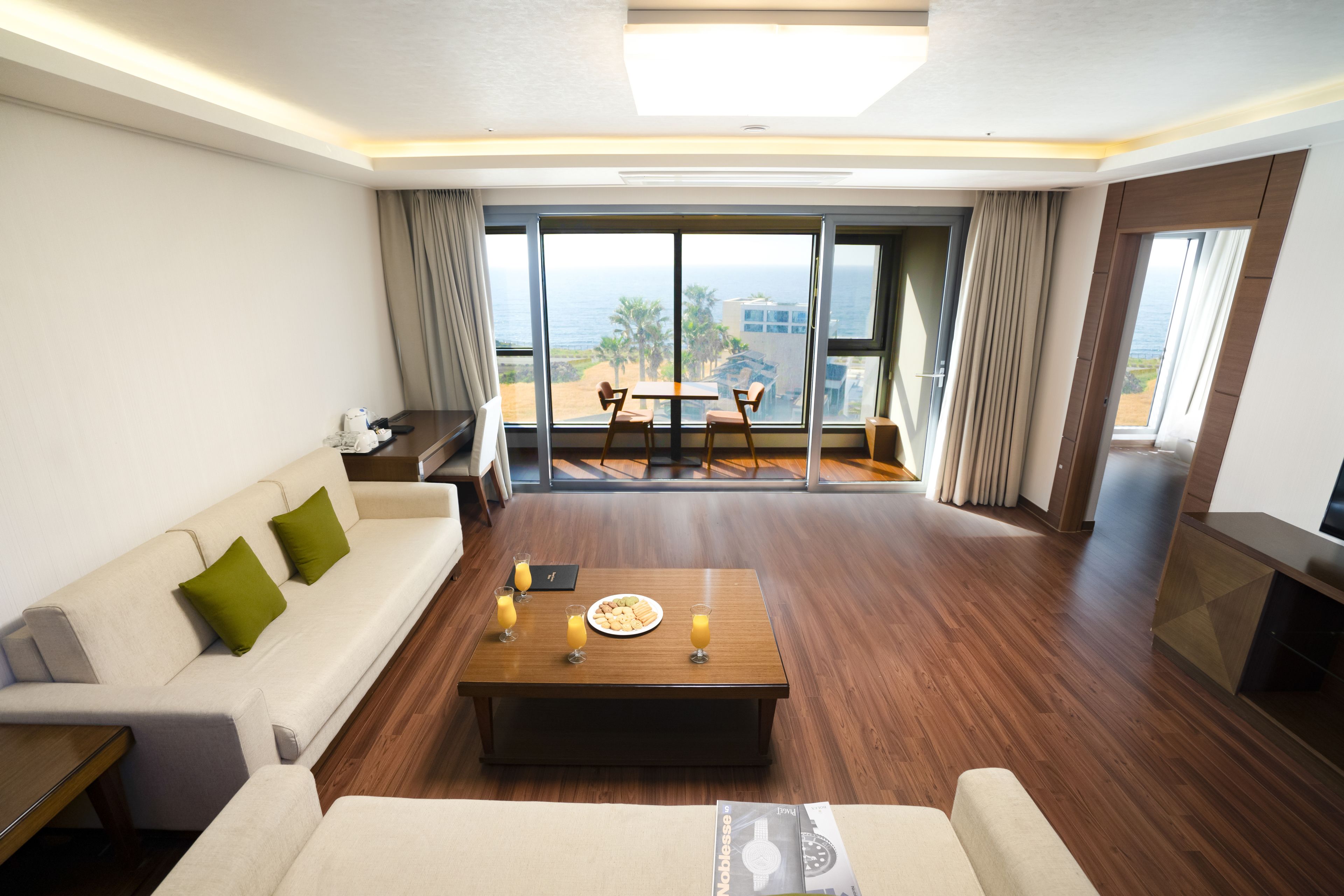 A modern hotel room with a sofa, a coffee table with drinks, a dining area with a table and chairs, and a large window with a view of the beach and ocean.