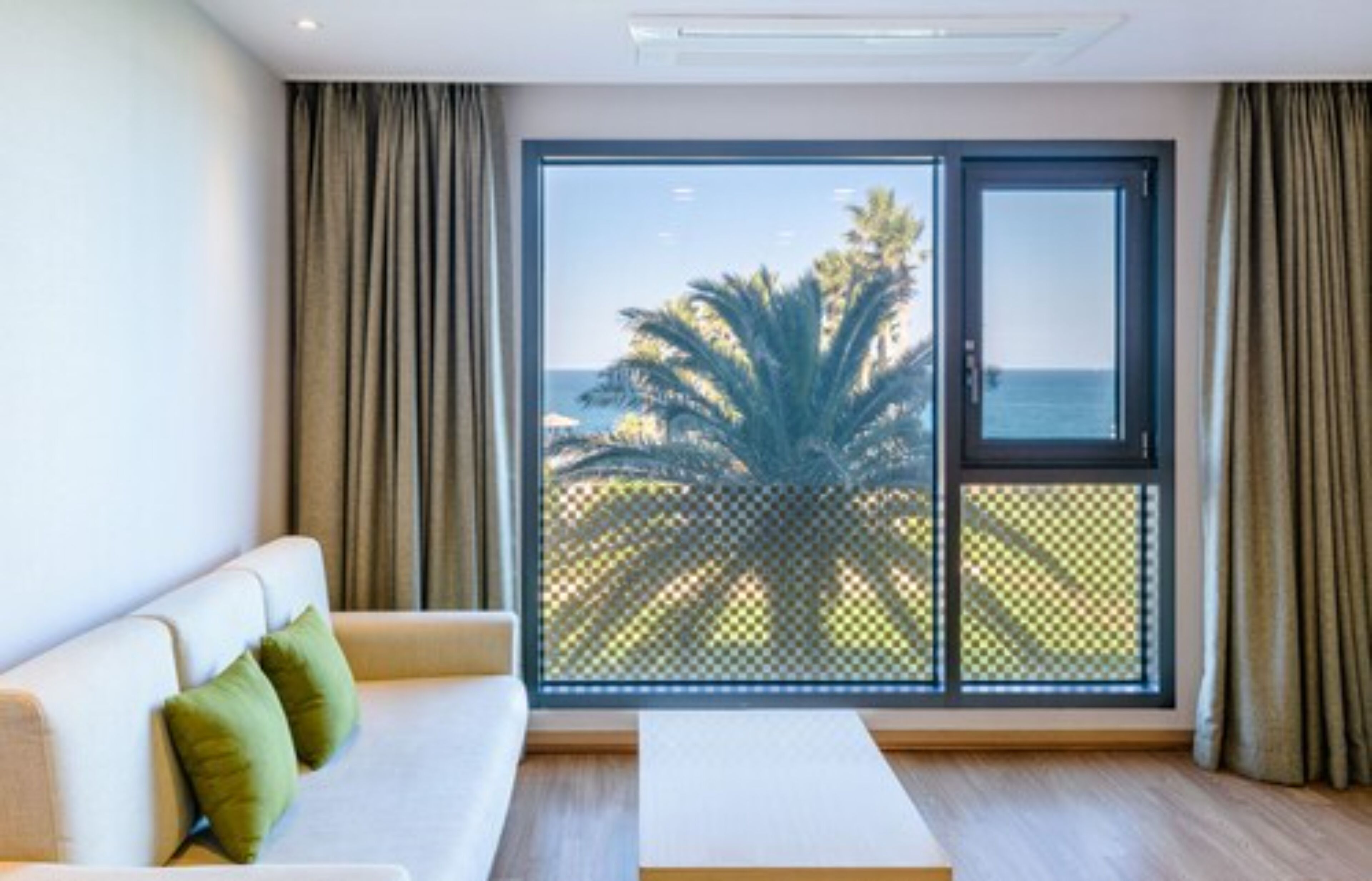 A modern room with a sofa, a small table, and a large window with a view of palm trees and the sea.