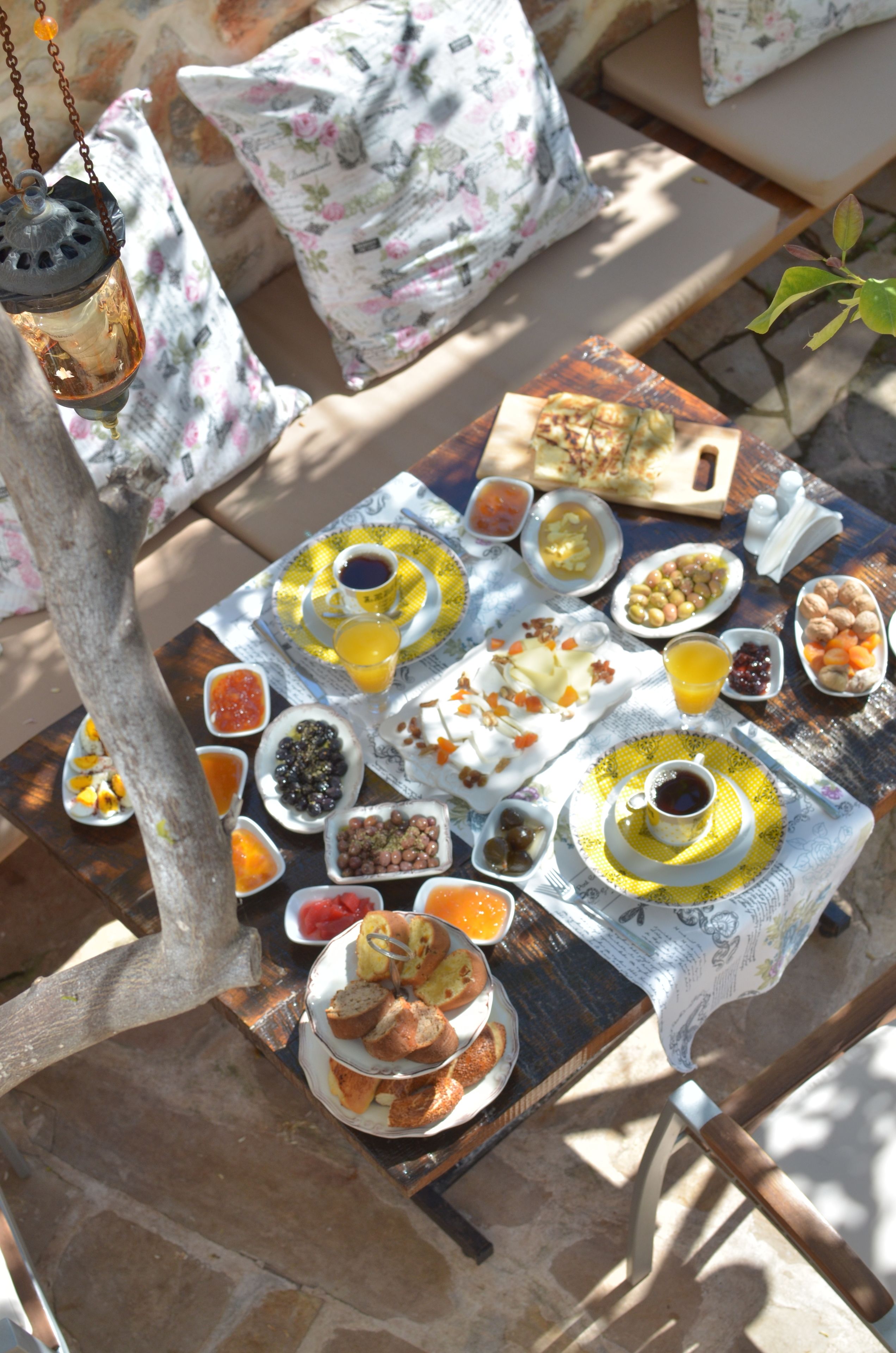 Mesa posta com diversos itens de café da manhã, incluindo pão, frutas e bebidas, rodeada por almofadas estampadas com flores e um lanterna pendente.