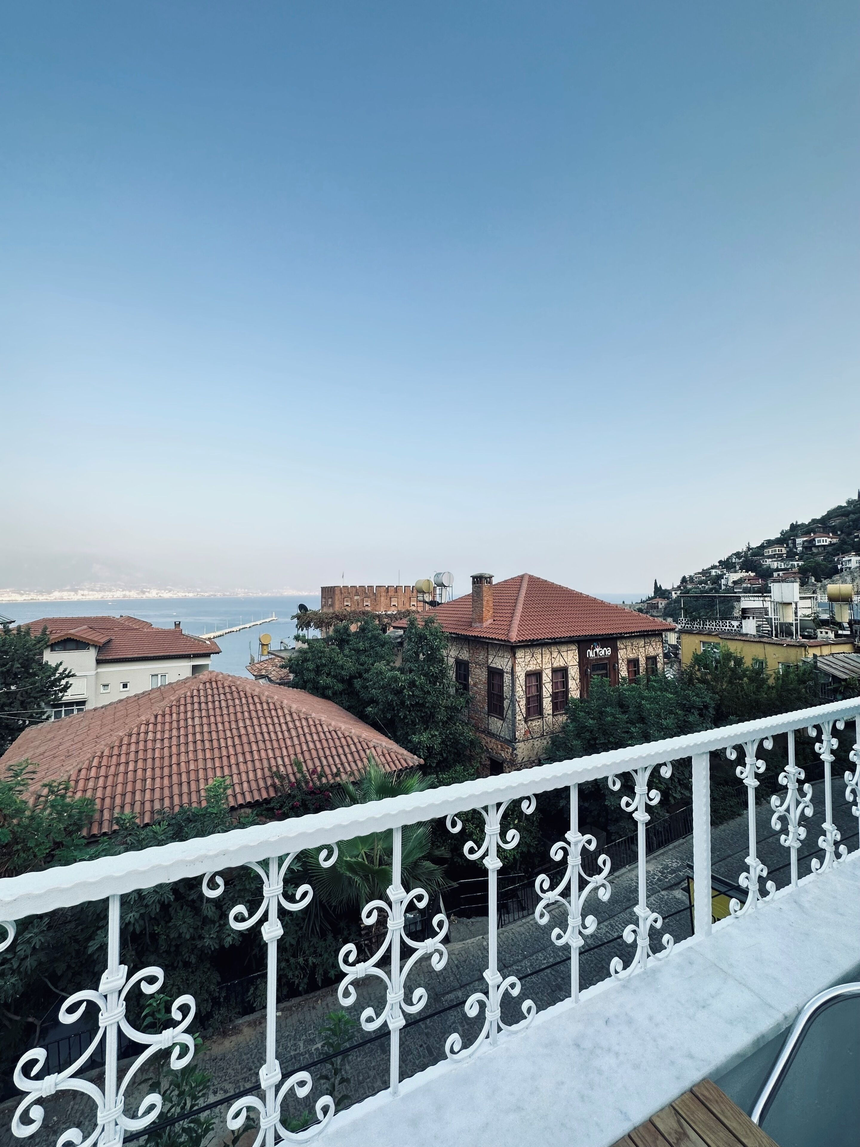 A balcony with white wrought iron railings overlooking a coastal town with terracotta-roofed buildings and a clear sky.