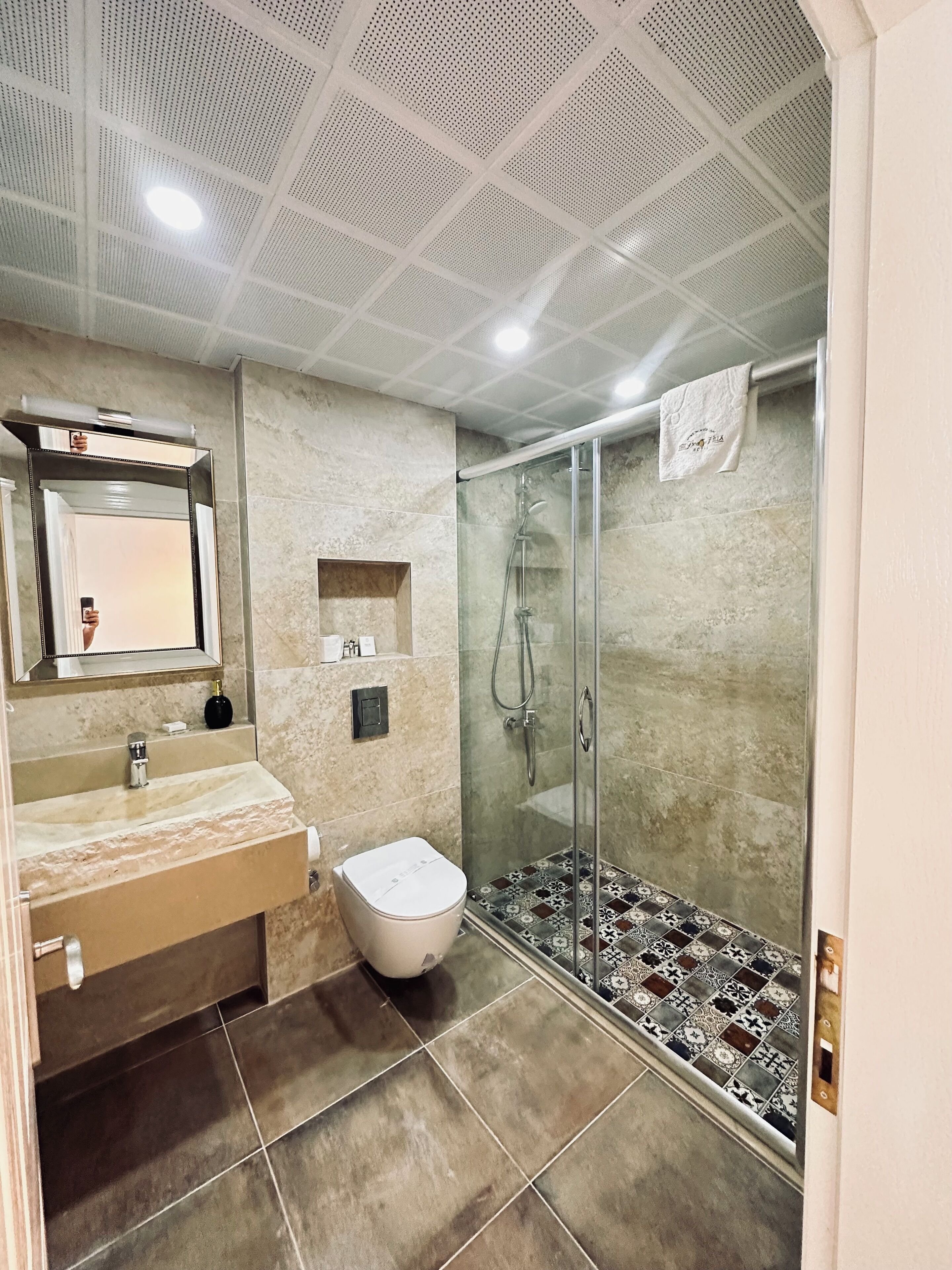 Een moderne badkamer met een marmeren wastafel, een toilet, een douche met glazen wanden en een plafond met een geperforeerd ontwerp.