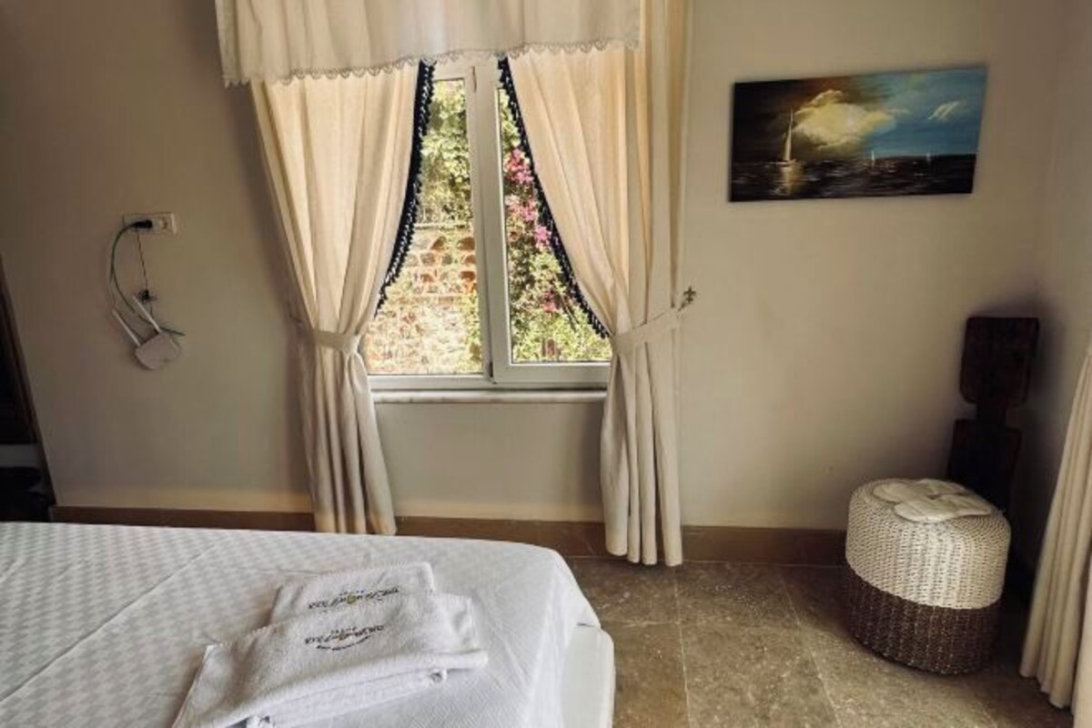 A hotel room with a bed, a window with curtains, a painting on the wall, and a small wicker ottoman.