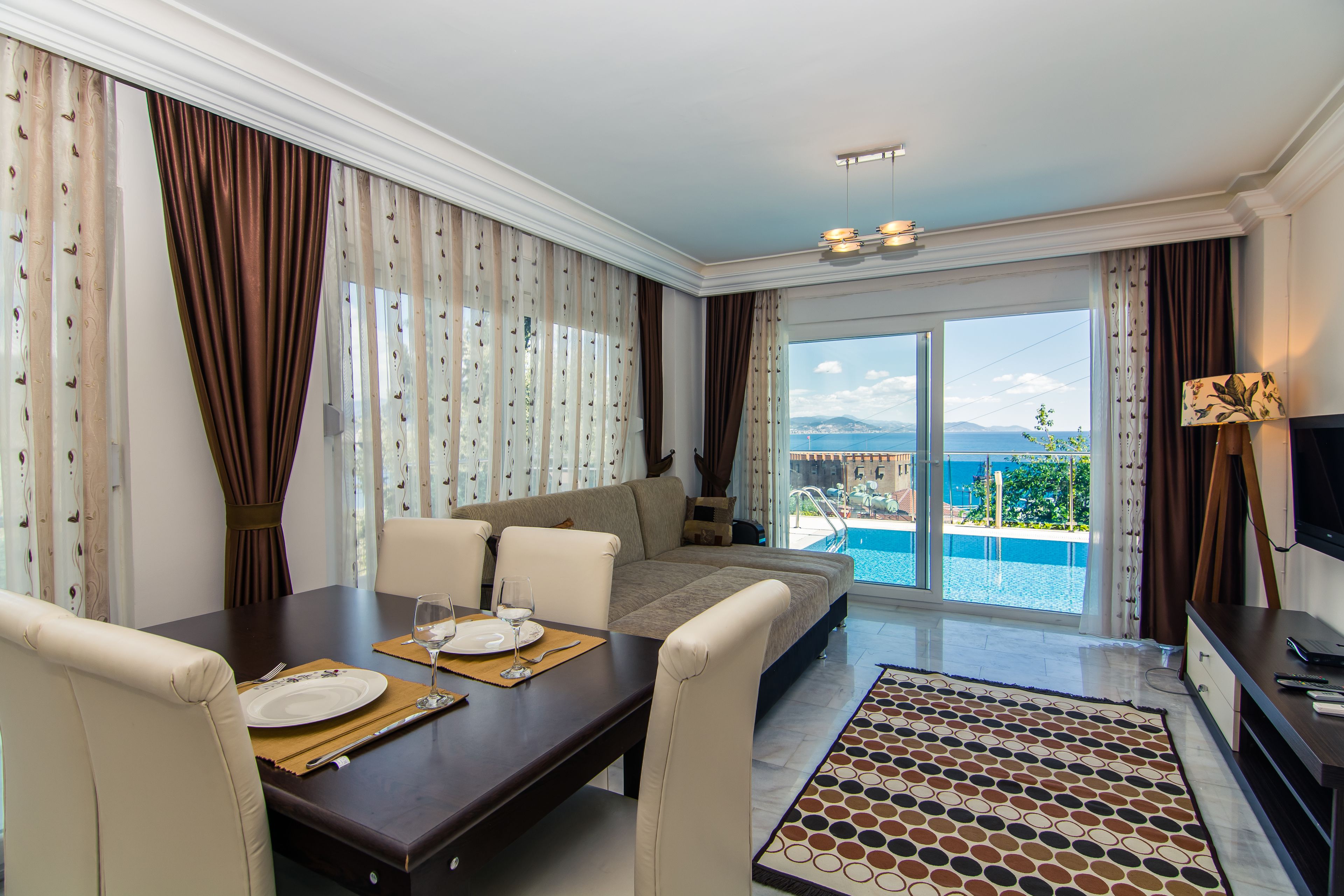 A modern hotel room with a dining area, a sofa, and a view of the pool and sea.