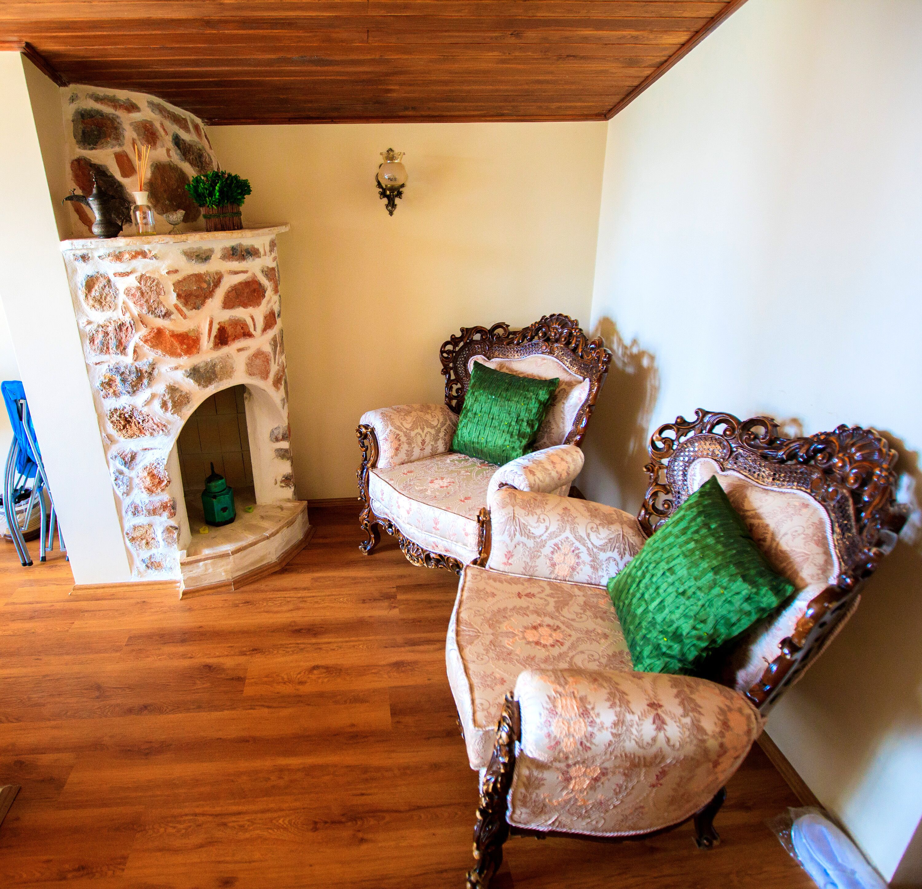 A room with a stone fireplace, two ornate armchairs with green cushions, and a wooden ceiling.