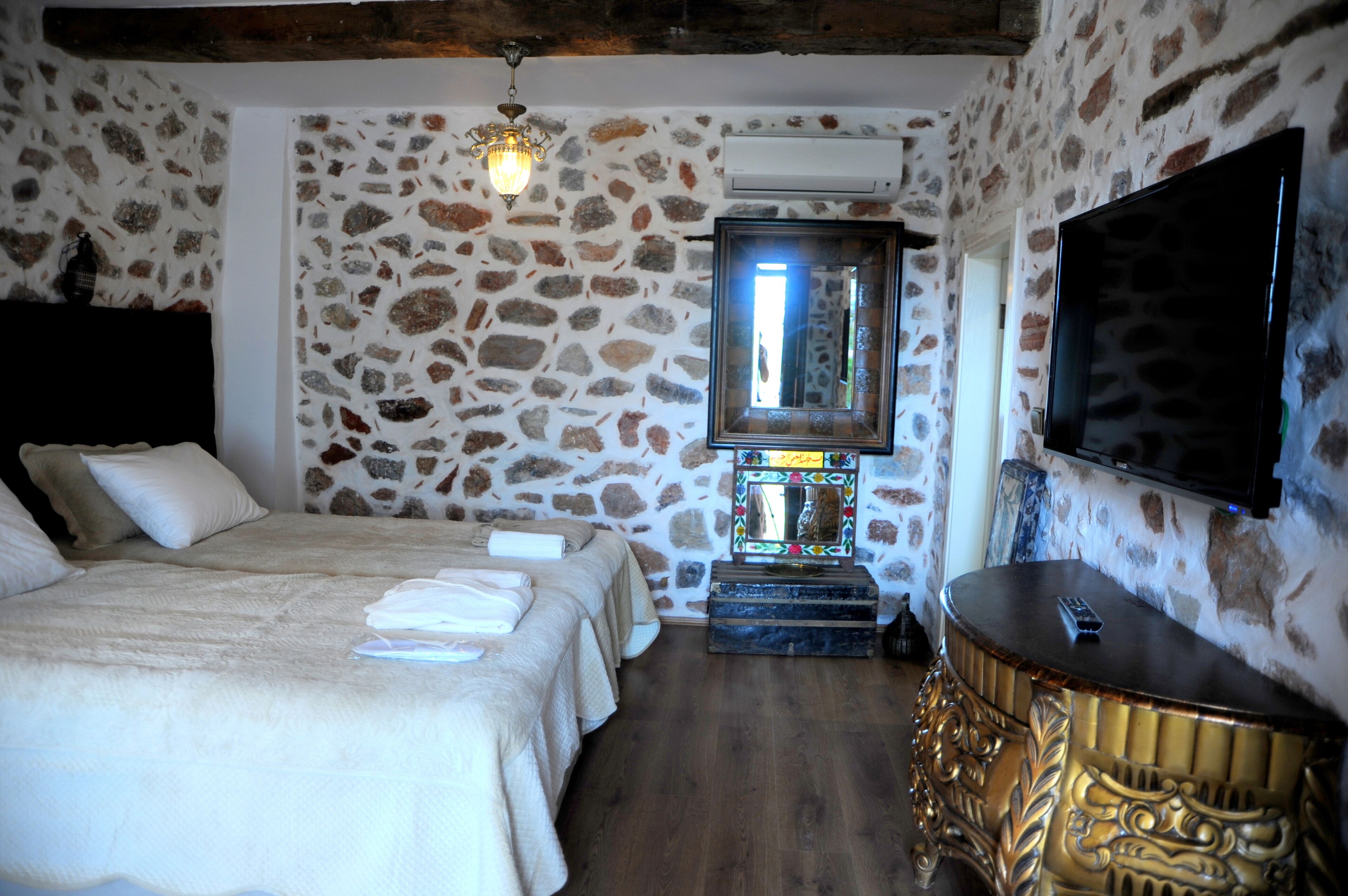 A bedroom with a stone wall, a bed with white linens, a wooden headboard, a bedside table with a remote control, and a TV mounted on the wall.