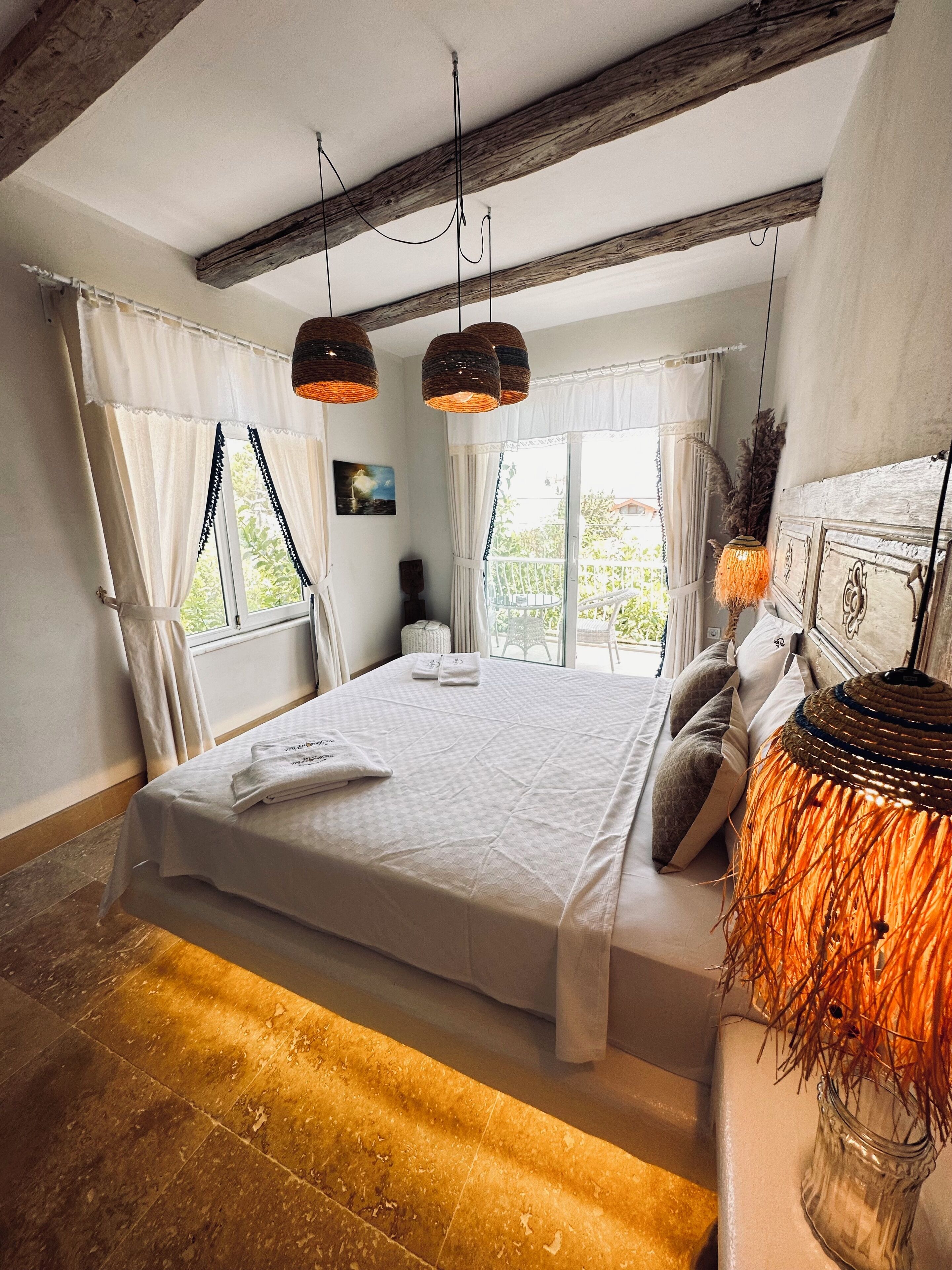 Un dormitorio con una cama grande, vigas de madera, una lámpara colgante y vistas al verde exterior.